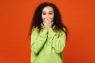 Young shocked surprised happy amazed woman of African American ethnicity wear green hoody casual clothes look camera cover mouth with hand isolated on plain red orange background. Lifestyle concept.