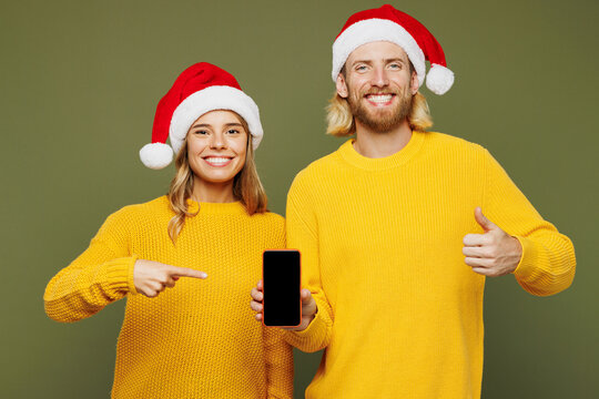 Merry young couple two friend man woman wear sweater Santa hat posing hold use blank screen mobile cell phone show thumb up isolated on plain green background Happy New Year Christmas holiday concept