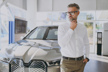 Adult confident minded man customer male buyer client wear shirt look aside prop up chin choose auto want to buy new automobile in car showroom vehicle salon dealership store motor show Sales concept