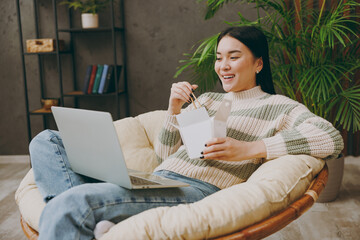 Young IT woman wear casual clothes sits on armchair hold use work on laptop pc computer eat Chinese food stay at home hotel flat rest relax spend free time in grey living room indoor. Lounge concept.