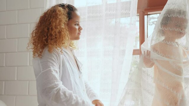 Cheerful mother playing with daughter hiding behind the curtains in living room at home. Little cute girl tease and enjoy with mother in bedroom at house. Family relationship concept.