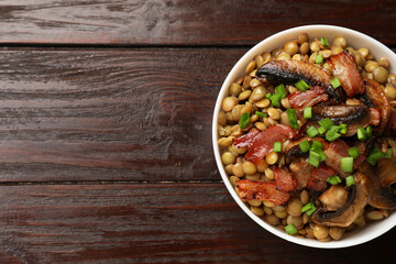 Delicious lentils with mushrooms, bacon and green onion in bowl on wooden table, top view. Space...
