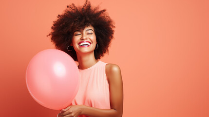 Joyful woman with an afro hairstyle, laughing and holding a pink balloon, wearing a light pink dress against a peach-colored background.