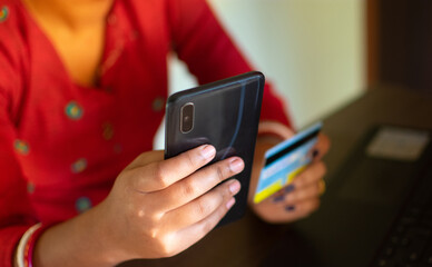 Shot of beautiful glad young female types number of credit card on smart phone, makes purchase online or checks bank account.