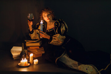 Young adult woman dressed in a medieval dress sitting near ancient books and candle light with...