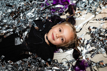 Cute happy smiling little girl in black dress lying on silver festive tinsel. Top view, emotions....