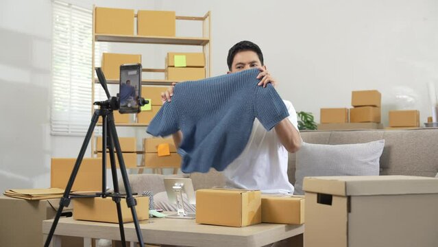 Asian people man happily displaying products to smartphone camera for live stream in living room with boxes, home office. clothing store