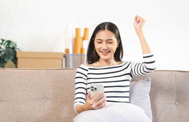 young Asian woman confidently using a smartphone, texting, chat with friends and video call sitting on a beige couch with a white cushion.