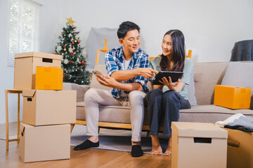 Asian couple is unpacking boxes in a festive living room with a Christmas tree, showing surprise and happiness.