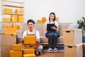 Asian man and woman in aprons work together, looking at paperwork and a laptop in a room filled with boxes.