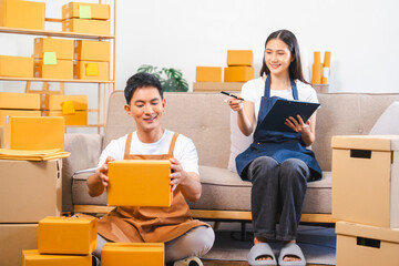 Asian man and woman in aprons work together, looking at paperwork and a laptop in a room filled with boxes.