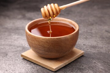 Pouring delicious honey from dipper into bowl on grey textured table, closeup