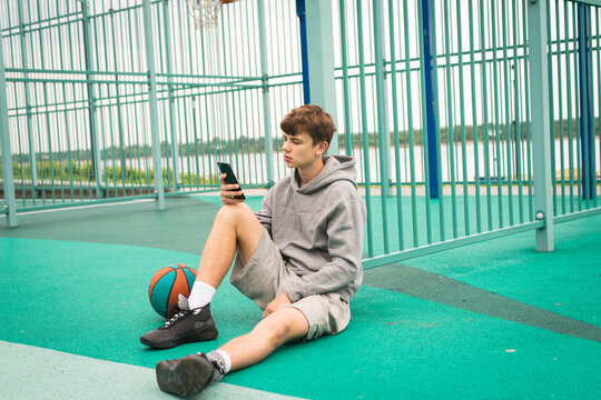 Teenage boy basketball player sitting alone at sportground, using smartphone