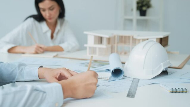 Professional architect engineer drawing blueprint and working together with young beautiful caucasian coworker on meeting table with safety helmet, house model, blueprint scatter around. Immaculate.