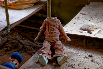 A headless doll with red overalls sitting on a dusty surface. Childrens doll without a head. A dirty, broken children's toy from a radiation-contaminated kindergarten in Chernobyl. - obrazy, fototapety, plakaty