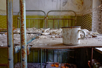 A white mug with a rusted rim and handle on a table. A white metal mug on a bed in an abandoned building in Chernobyl.