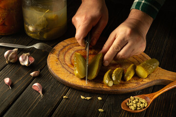 Close-up of a man hands using a knife to cut a pickled cucumber on the kitchen table for preparing...