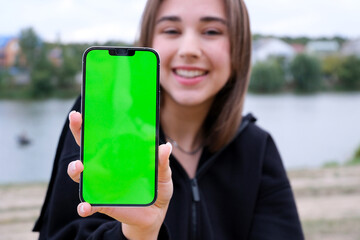 young cheerful girl against backdrop of the lake points to phone screen of smartphone. finger smiles laughs Good advertising for walks travel fun girl with fair skin in black clothes beautiful teeth