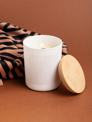 Handmade candle in white cup with wooden lid on brown background. Candle and fabric with animal print