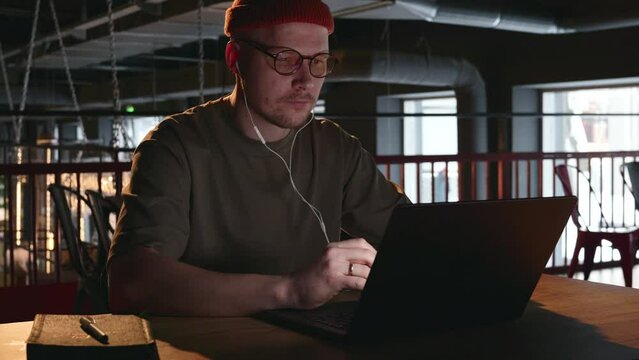 A young remote worker, a man of 30 years old, works in a coworking behind laptops and sticks headphones in his ears to listen to music. The man sticks in the headphones to evaluate the song sent. 4k