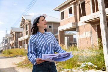 Caucasian woman enginee wears helmet and reflective safety uniform holding blueprints walking to inspect construction work in housing site.