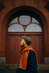 Naklejka premium A caucasian man with a beard standing in front of an old red wooden door wearing an orange scarf and a blue coat looking away from the camera.