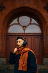 A caucasian man with a beard wearing wireless headphones using a phone standing in front of an old red wooden door wearing an orange scarf and a blue coat looking to the side.