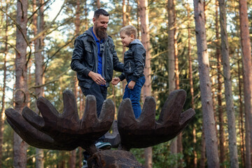 A happy father and son stand tall on a wooden moose sculpture in the antler forest