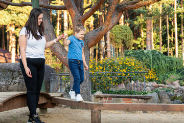A brave, athletic boy travels on a wooden beam with ease and is slightly held by his mother's hand.