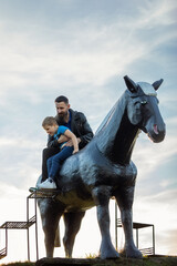 Portrait of a father and his little son on a big black horse against the background of the sky. Dad...
