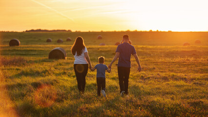 A young family from the back walking through the wide country fields holding hands in the rays of...