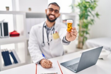 Young hispanic man doctor holding pills bottle writing on document at clinic