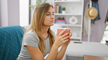 Attractive young blonde woman comfortably sitting indoors in her apartment's living room, enriching her morning by relaxing and drinking a warm espresso coffee while doubtingly pondering.