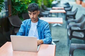 Young hispanic man using laptop sitting on table at coffee shop terrace