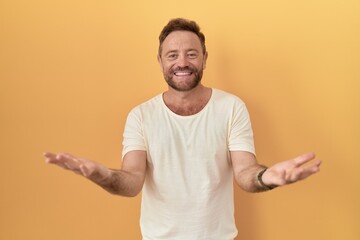 Middle age man with beard standing over yellow background smiling cheerful with open arms as friendly welcome, positive and confident greetings