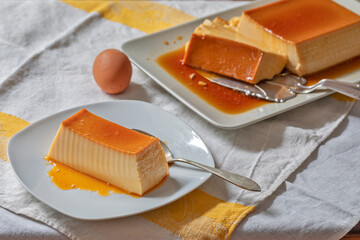 In the foreground, a slice of homemade creme caramel on a small plate with a teaspoon. Behind an...