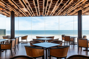 Contemporary waterfront cafe interior with ocean panorama through floor-to-ceiling windows, surrounded by wooden tables and chairs. Leisure and travel.