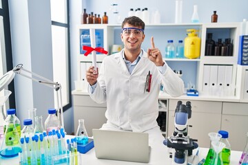 Young hispanic man working at scientist laboratory holding diploma surprised with an idea or question pointing finger with happy face, number one
