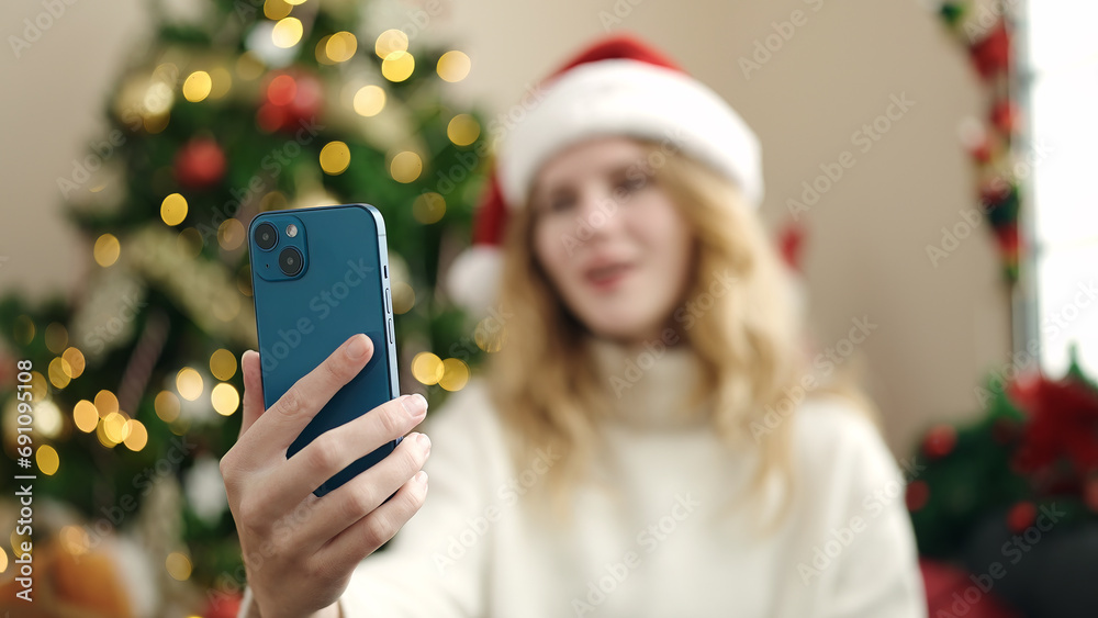 Sticker Young blonde woman making selfie by smartphone sitting on sofa by christmas tree at home