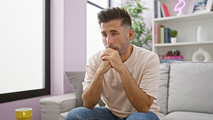 Pensive young hispanic man seeking comfort on sofa, close-up indoor portrait highlighting the...