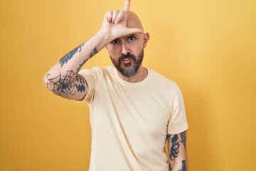 Hispanic man with tattoos standing over yellow background making fun of people with fingers on...