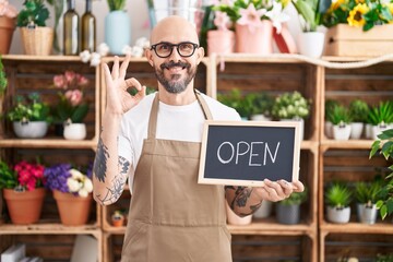 Hispanic man with tattoos working at florist holding open sign doing ok sign with fingers, smiling...