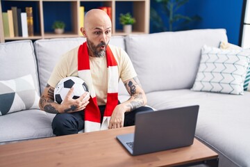 Hispanic man with tattoos watching football match hooligan holding ball on the laptop scared and...
