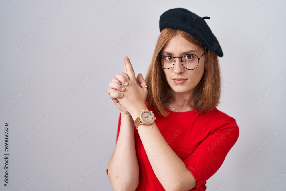 Poster young redhead woman standing wearing glasses and beret holding symbolic gun with hand gesture, playi