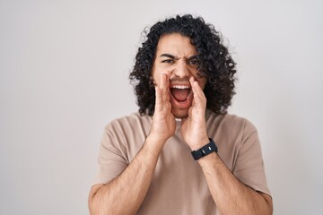Fototapeta na wymiar Hispanic man with curly hair standing over white background shouting angry out loud with hands over mouth