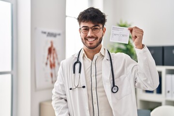 Hispanic doctor man with beard holding covid record card looking positive and happy standing and...