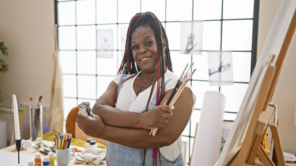 Confident african american woman artist with braids, paintbrushes in hand, standing with crossed...