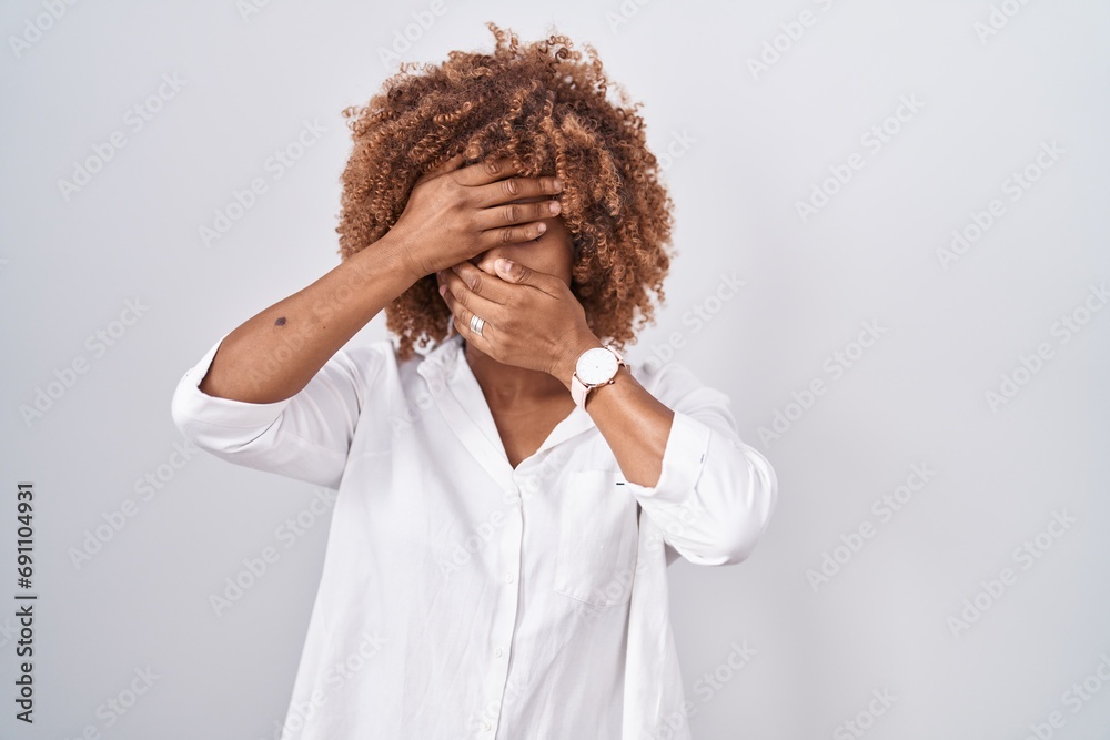 Sticker young hispanic woman with curly hair standing over white background covering eyes and mouth with han