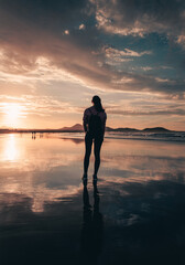 Vertical photo of free woman tourist on the beach during the sunset golden hour looking at the ocean. Woman on the beach with her reflection on wet sand - lifestyle concept.