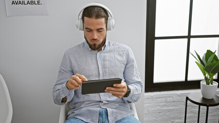 Young hispanic man using touchpad and headphones sitting on chair at waiting room